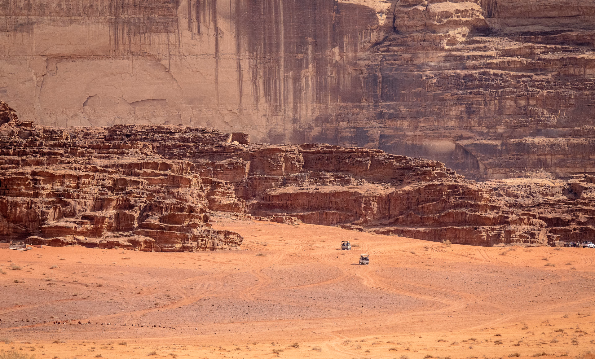 Two off-road vehicles crossing red desert sand toward dramatic sandstone cliffs in Wadi Rum